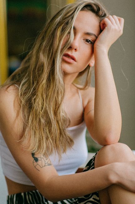 Portrait of a blonde woman with tattoos sitting and touching her forehead. Casual indoor setting.