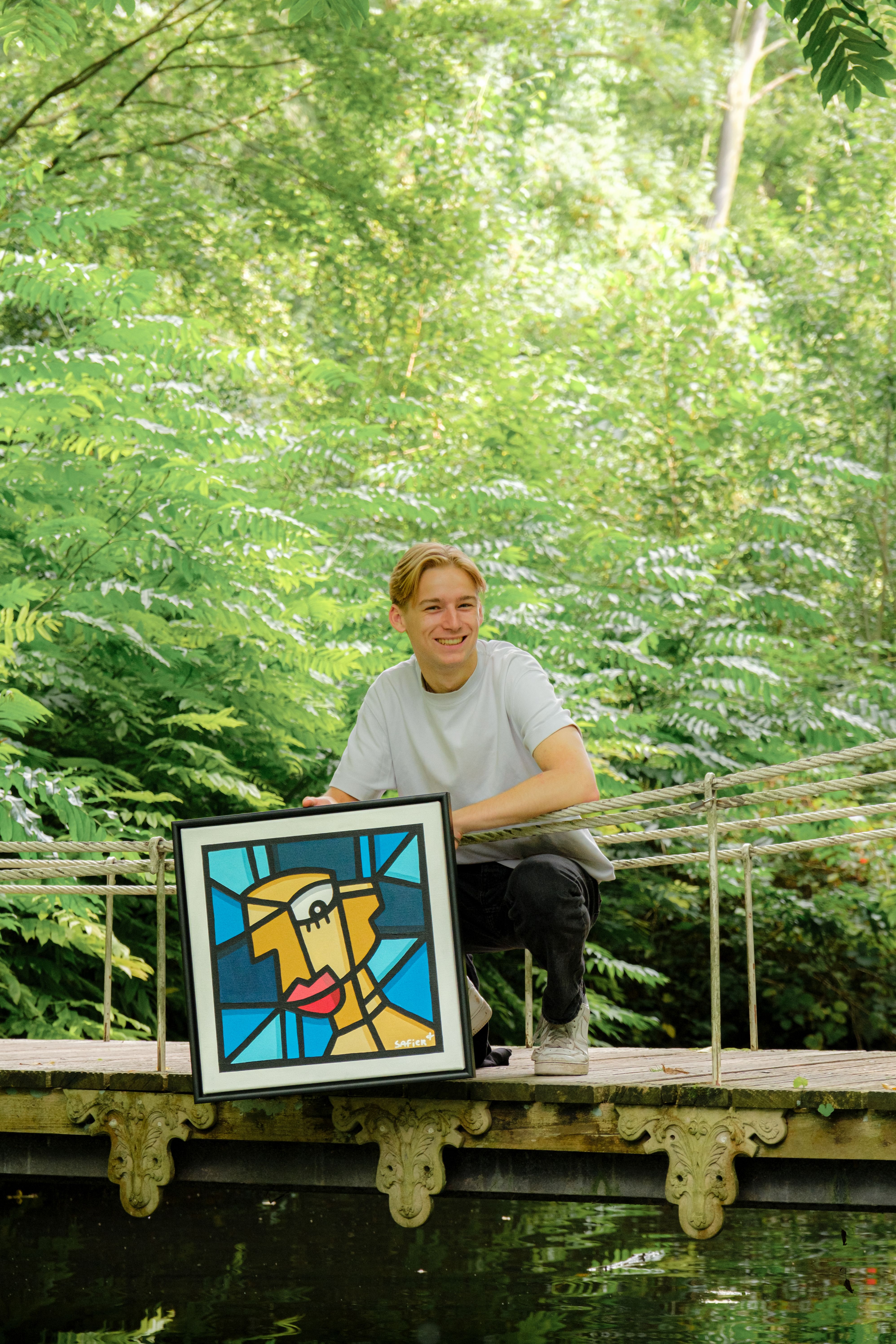 Smiling young man with framed cubist art on a forest bridge.
