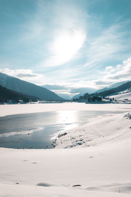 A serene snowy landscape featuring a sunlit frozen lake surrounded by mountains under a clear sky.