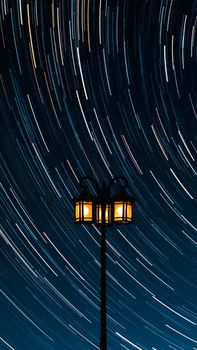 Vertical photo of star trails with an illuminated street lamp in the foreground at night.
