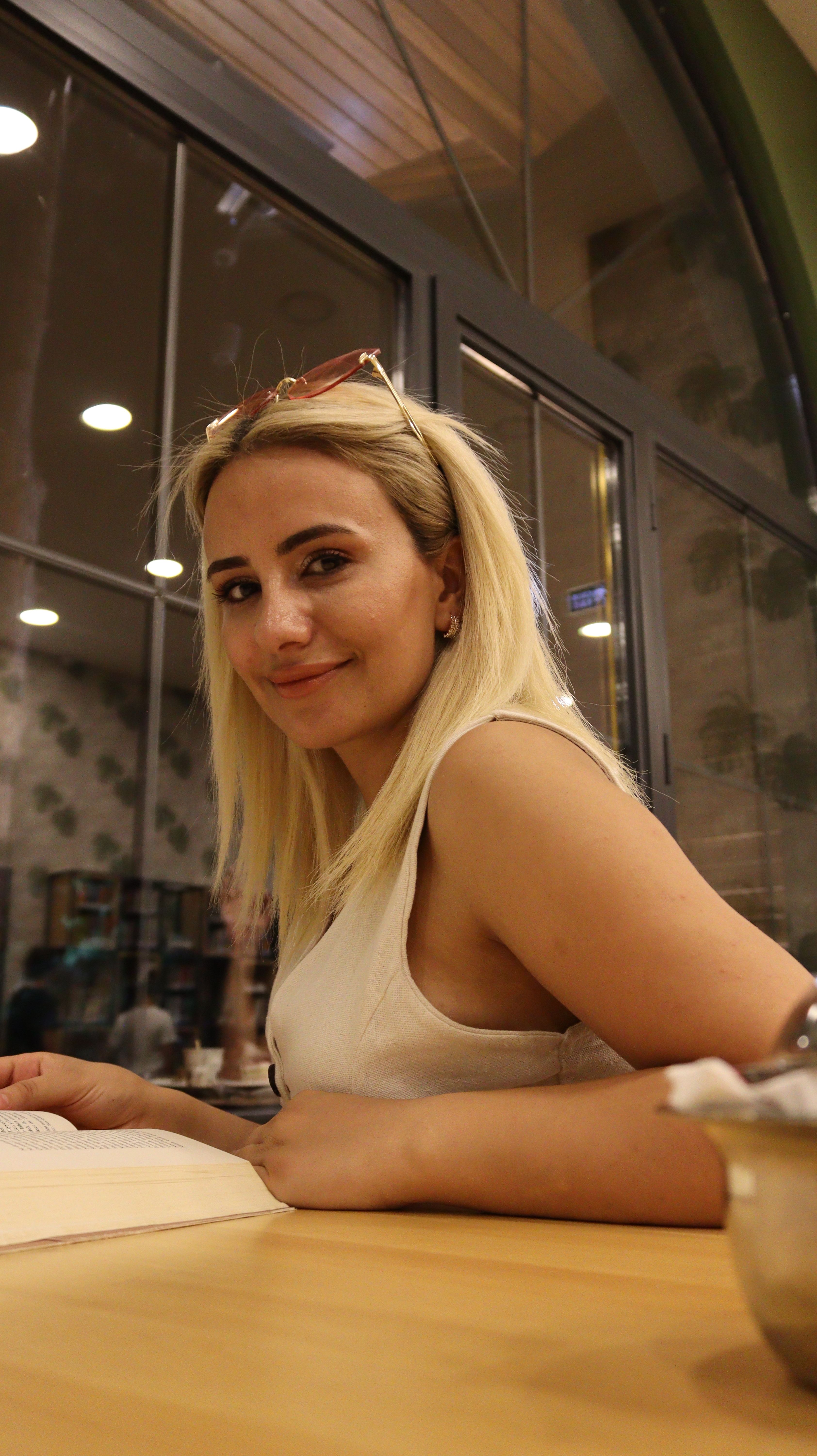 A young woman with blonde hair smiles while reading a book at a cozy café.