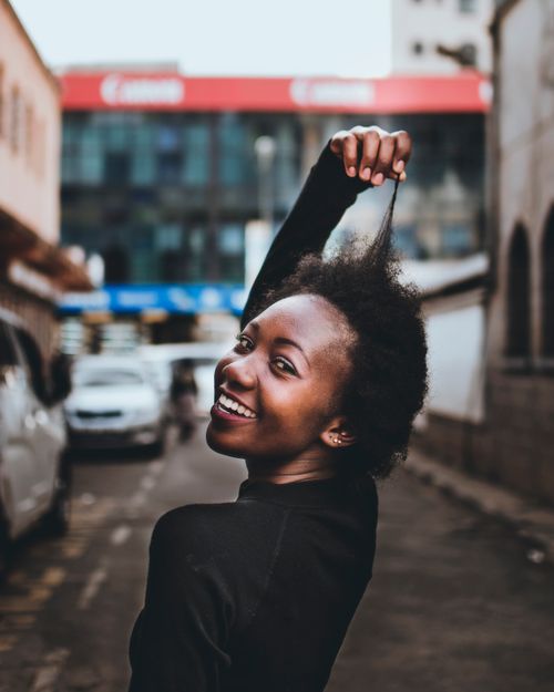 Free Black woman with curly hair smiling on a city street with a playful pose. Stock Photo