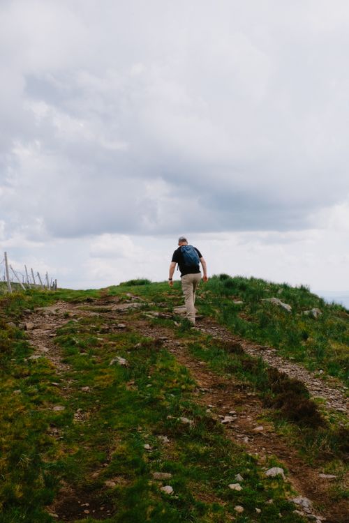 Free A man with a backpack walks up a rocky path on a hilltop, under a cloudy sky, epitomizing outdoor adventure. Stock Photo