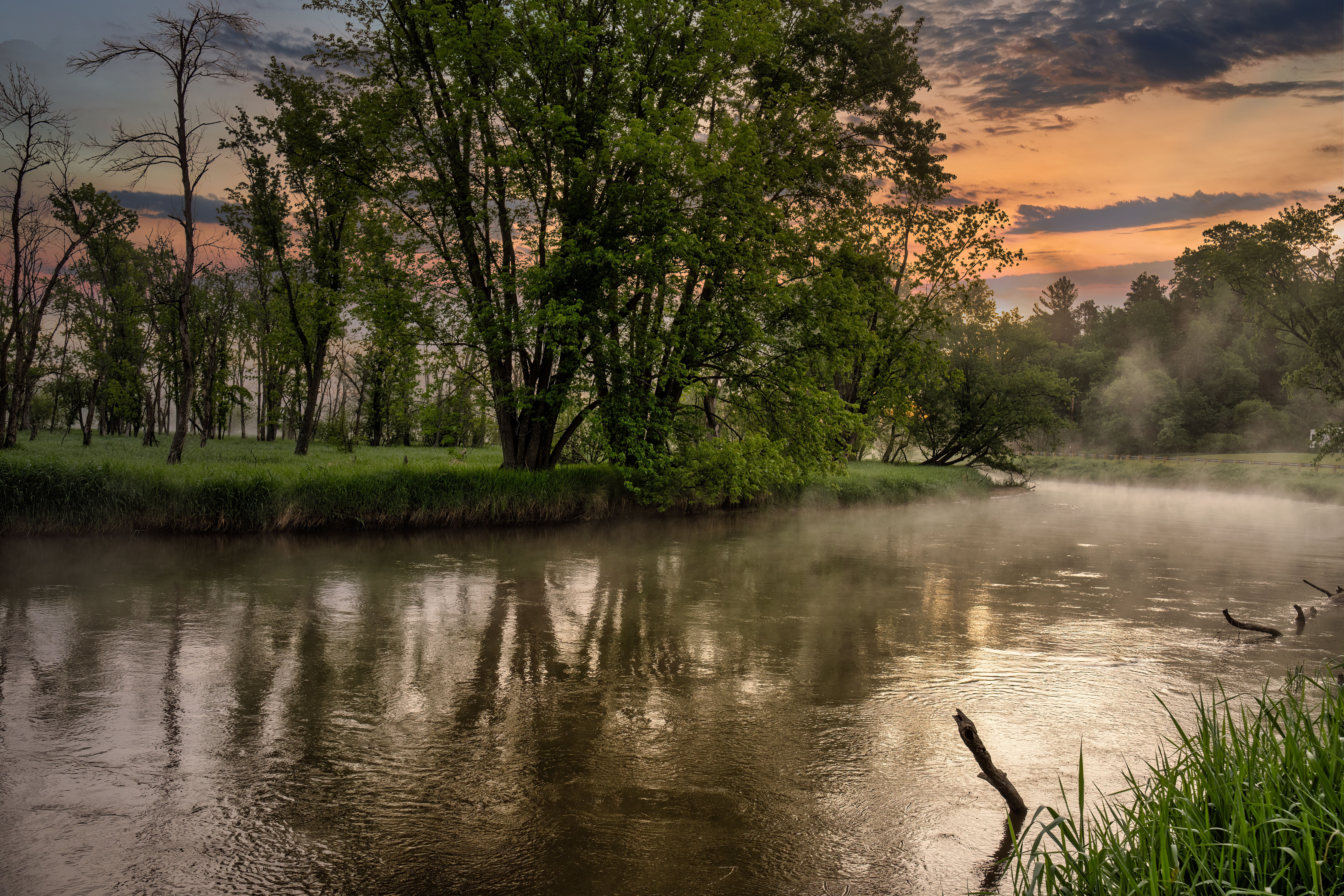 Tranquil river scene at sunset with mist and lush forest trees in serene wilderness.