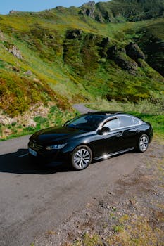 Sleek black car parked on a winding road amidst lush green mountains in bright daylight.