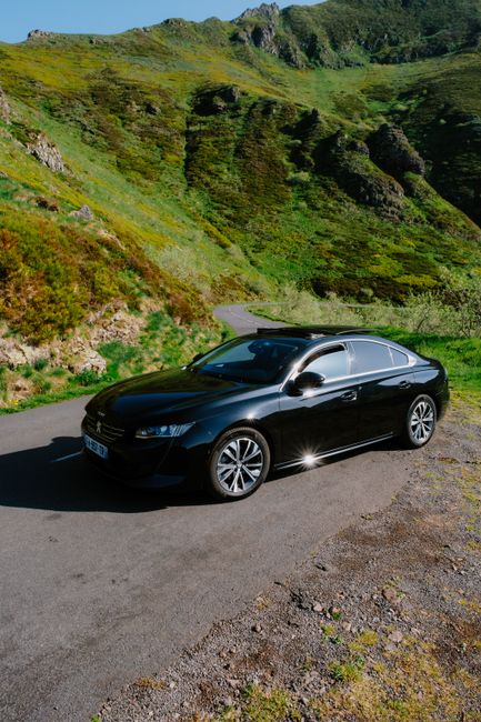 Sleek black car parked on a winding road amidst lush green mountains in bright daylight.