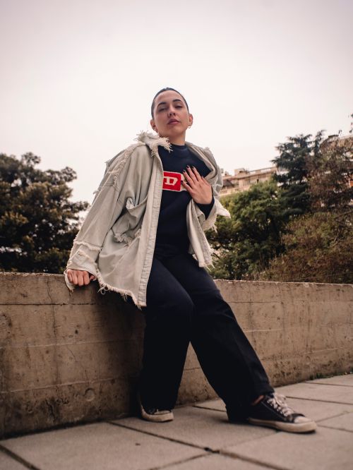 Free Stylish young woman sits on urban concrete wall, wearing black pants and gray jacket. Stock Photo