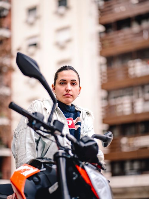 Free Confident woman in a white jacket posing with a motorcycle against a city backdrop. Stock Photo