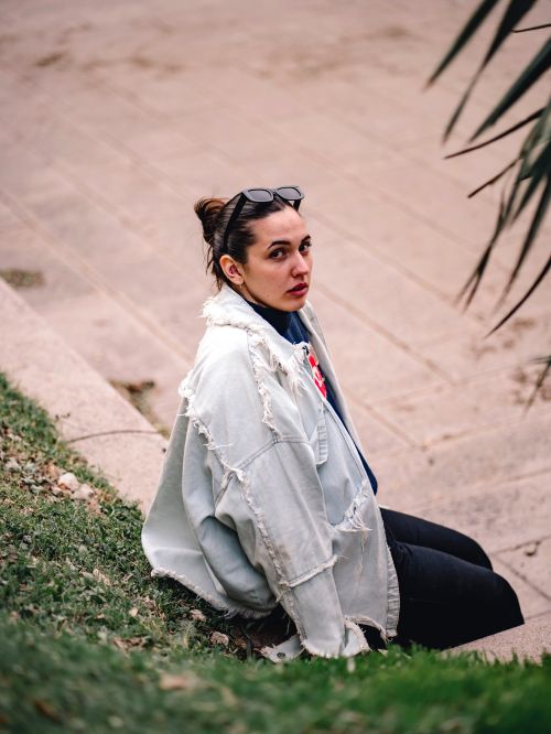 Free Woman in a denim jacket and sunglasses sitting on outdoor steps, embodying casual fashion. Stock Photo