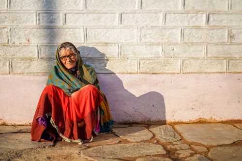 Free Portrait of an elderly woman sitting outdoors against a stone wall, wrapped in a colorful scarf. Stock Photo