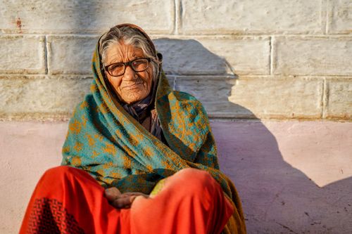 Free An elderly woman sits warmly wrapped in a colorful shawl against a brick wall, capturing serene outdoor moments. Stock Photo