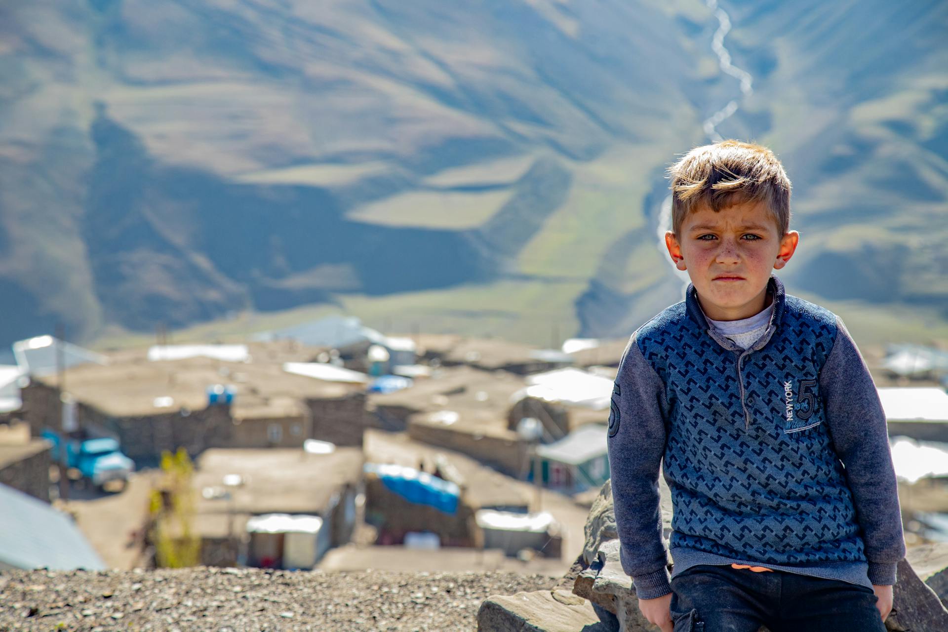 A young boy with short hair sitting outdoors in Khinalug, Azerbaijan with a scenic mountainous backdrop.