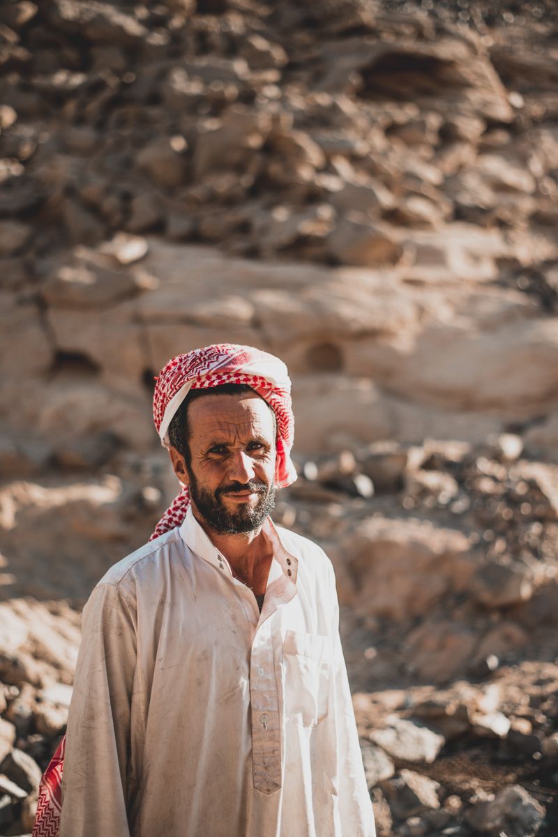 Portrait of a Middle Eastern man wearing a keffiyeh, standing in a rocky desert landscape.