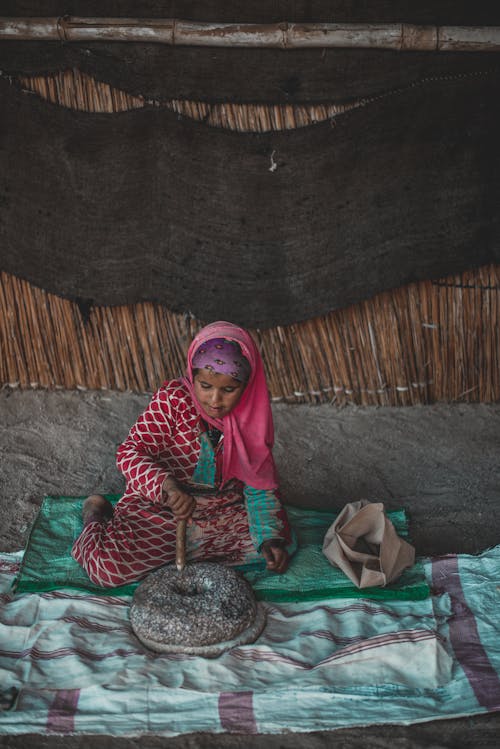 Full body of ethnic woman in traditional clothes sitting on floor with stone utensils in hut