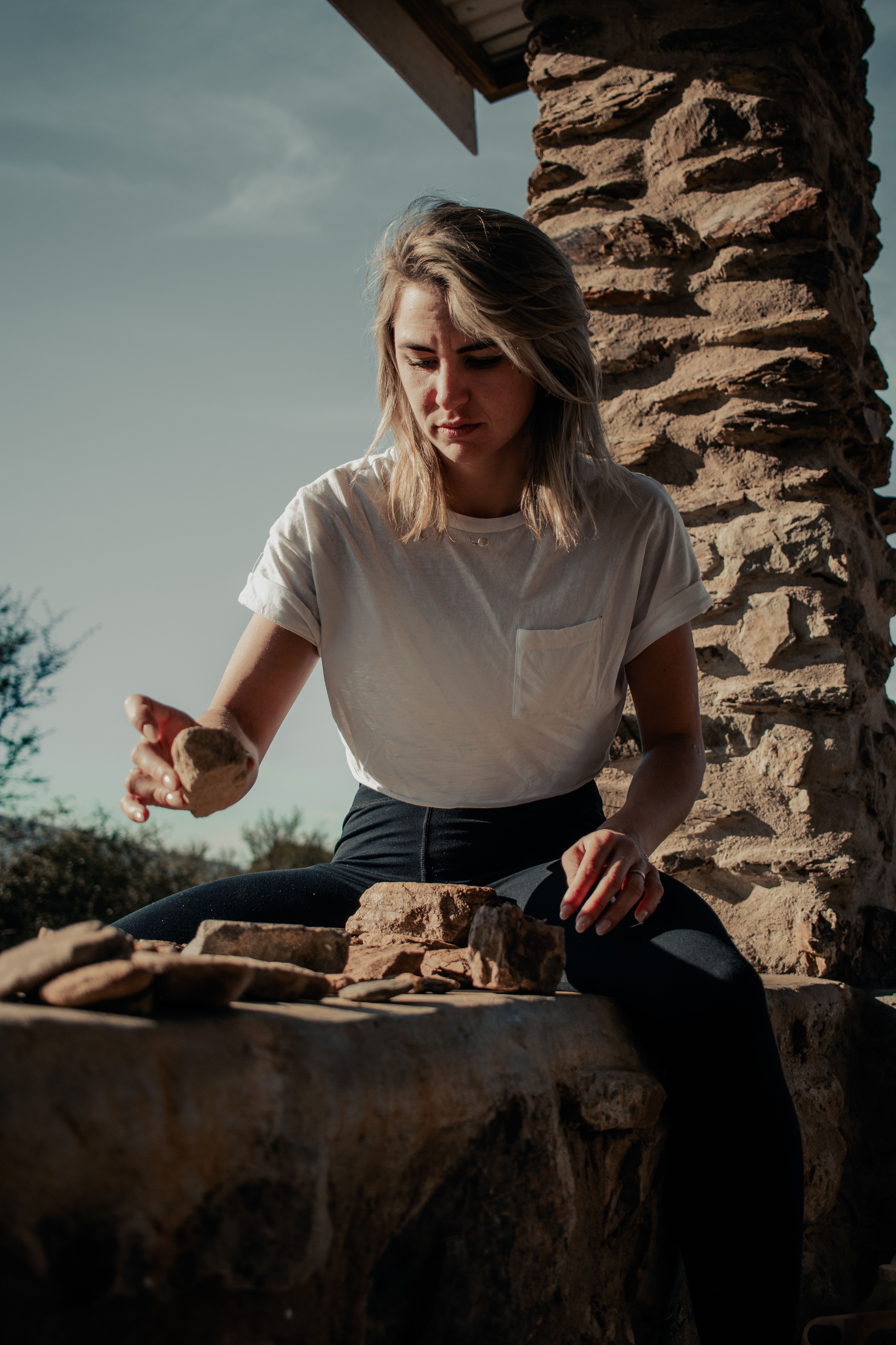 A woman in casual white top sits and arranges stones by a rustic wall in sunlight.