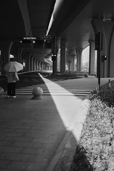 Black and white photo of a person under an umbrella at a city overpass.