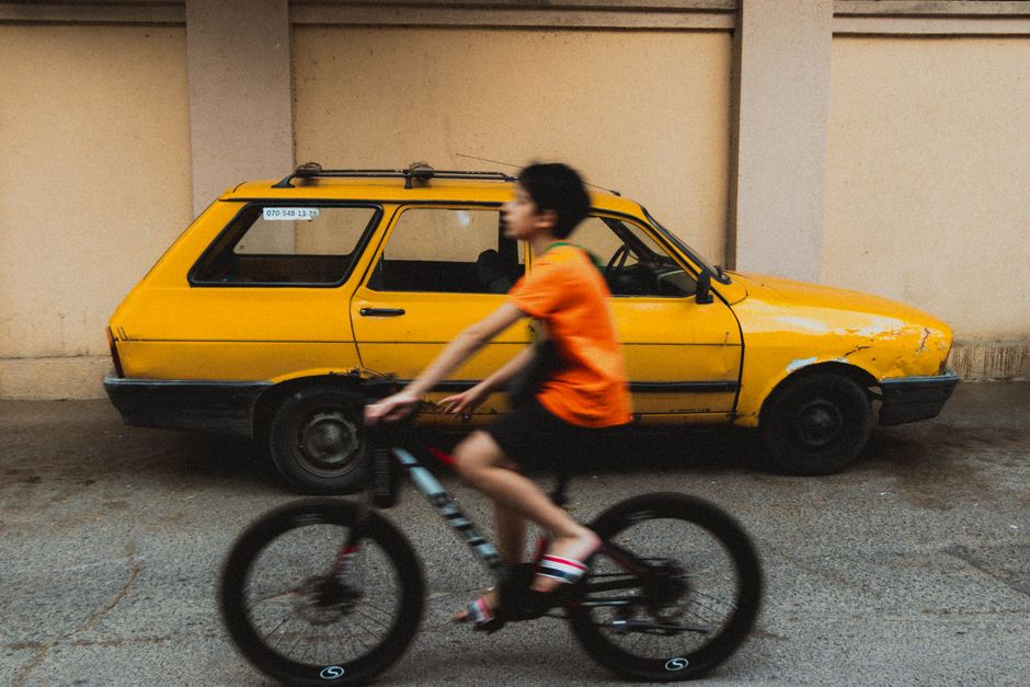 Blurry child cycling past a vintage yellow car, capturing urban street movement.