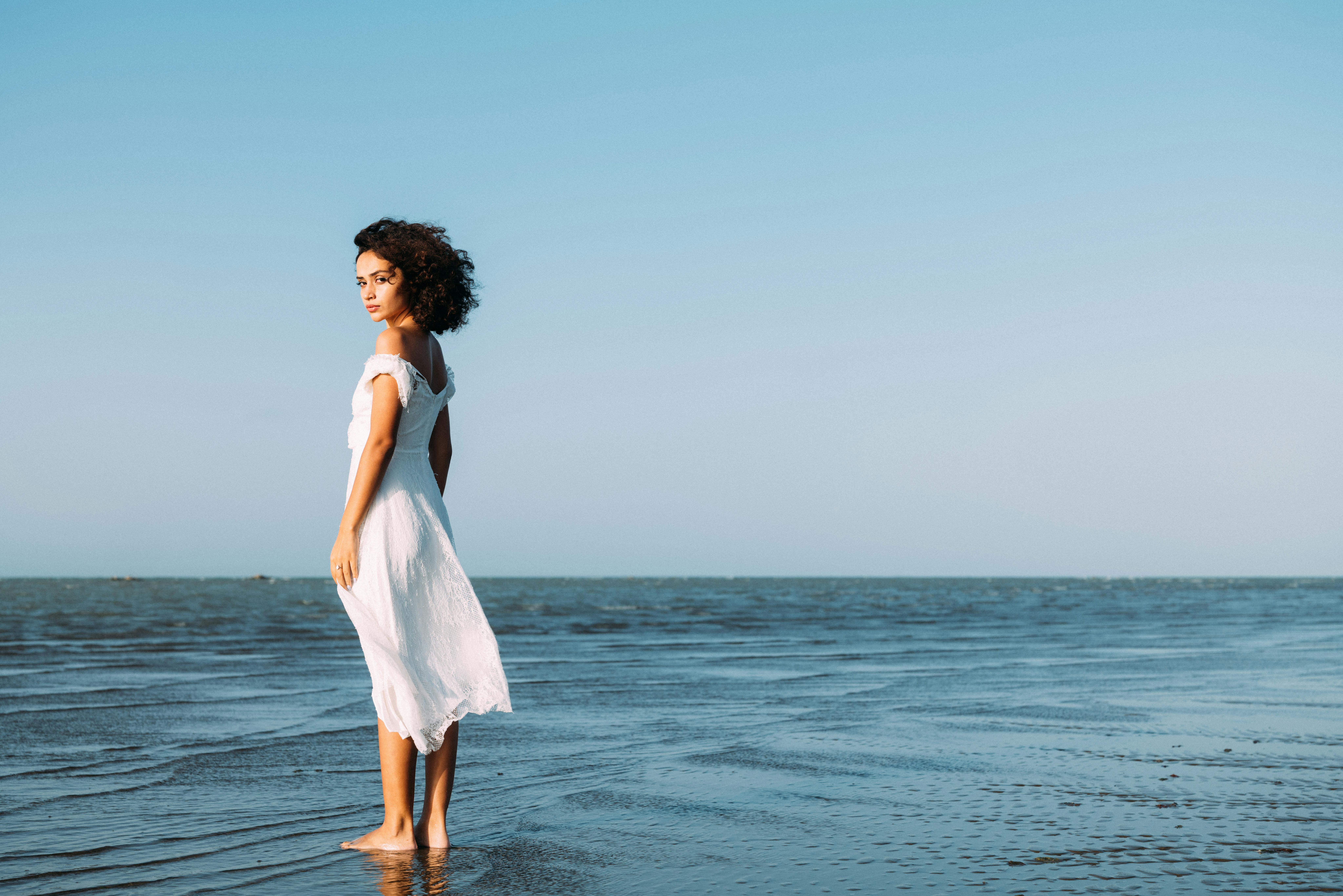 Young woman in a white dress standing on the beach with a serene ocean background.