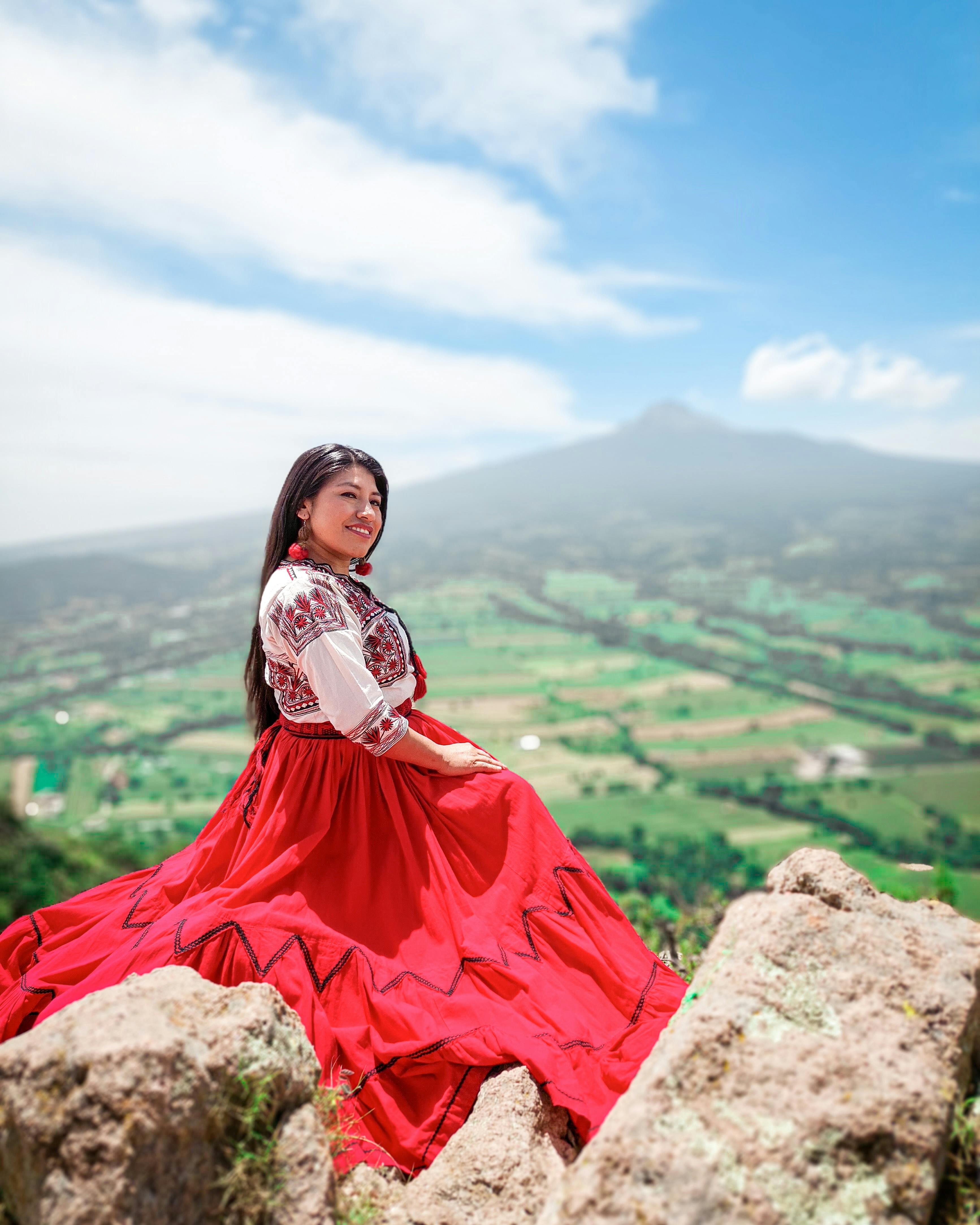 Portrait of a woman in vibrant traditional dress sitting on rocky terrain in Tlaxcala, Mexico, against a scenic backdrop.