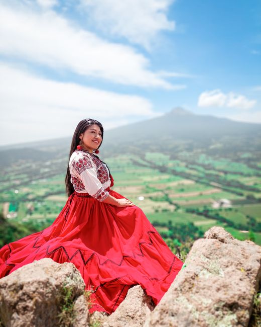 Portrait of a woman in vibrant traditional dress sitting on rocky terrain in Tlaxcala, Mexico, against a scenic backdrop.