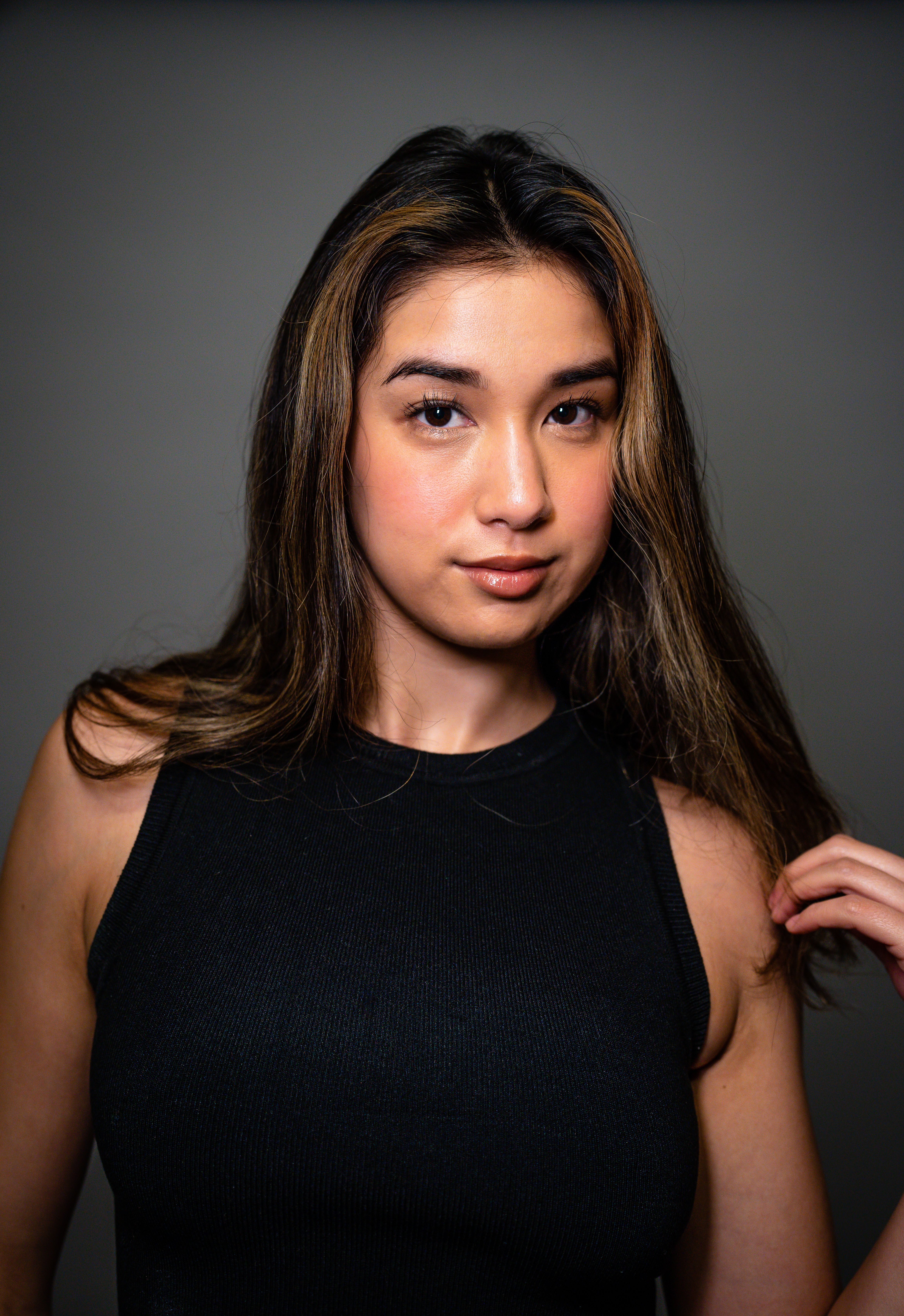 Portrait of a young woman with long brown hair wearing a black top against a gray background.