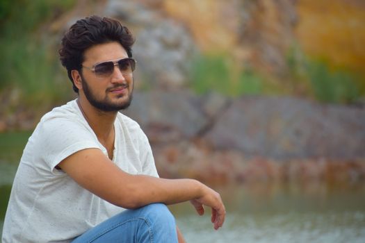 Outdoor portrait of a man wearing sunglasses and sitting by a scenic background, exuding a relaxed vibe.