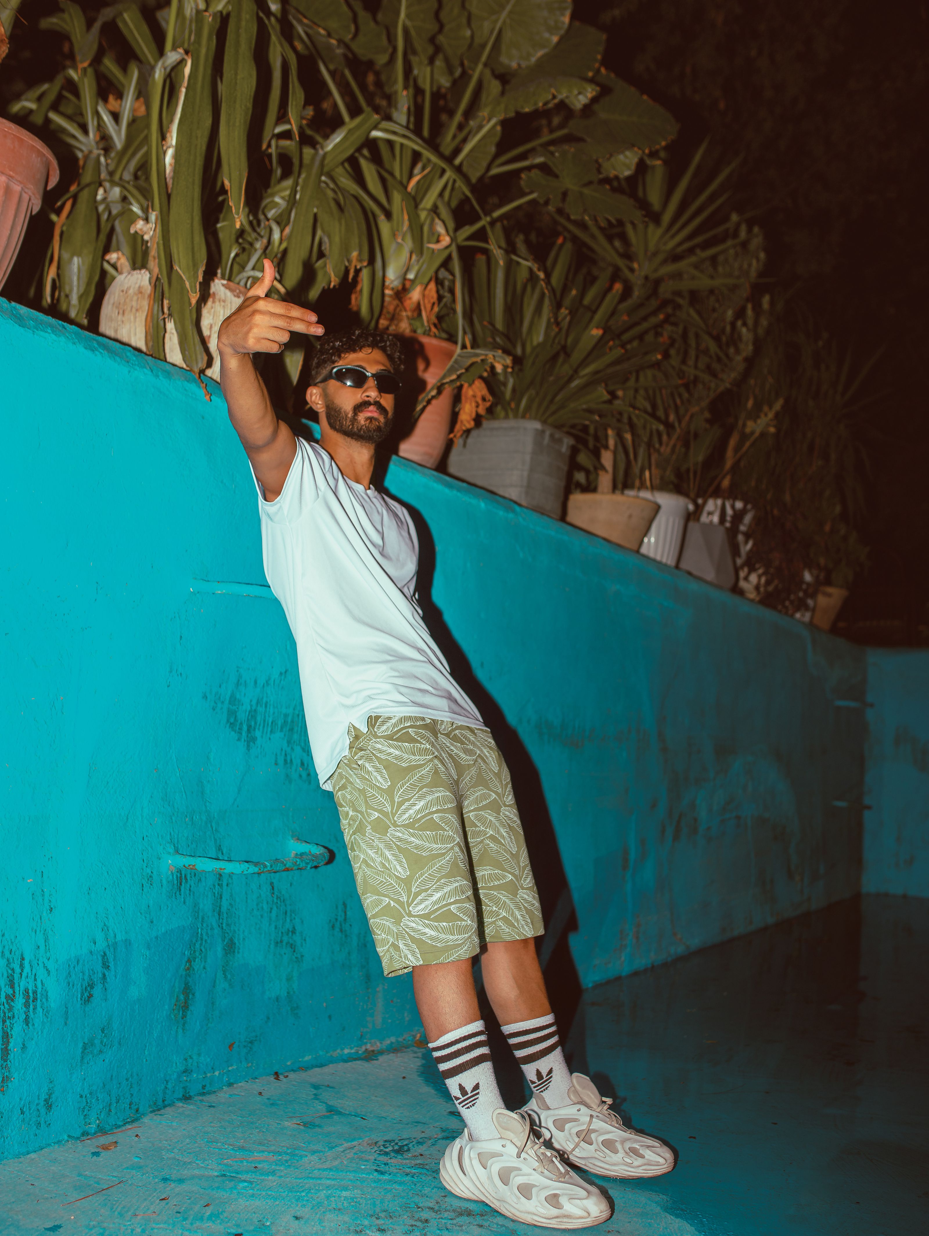 Homem fashion com barba e óculos de sol posando ao lado de uma parede azul à noite.
