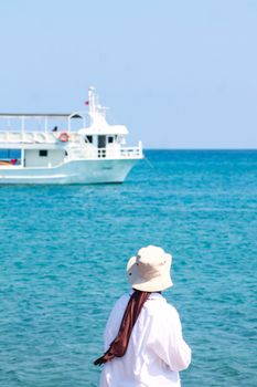 A woman in a hat stands on the shore with a boat in the sea.