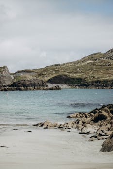Serene view of a rocky coastal landscape with calm sea and hills under a cloudy sky.