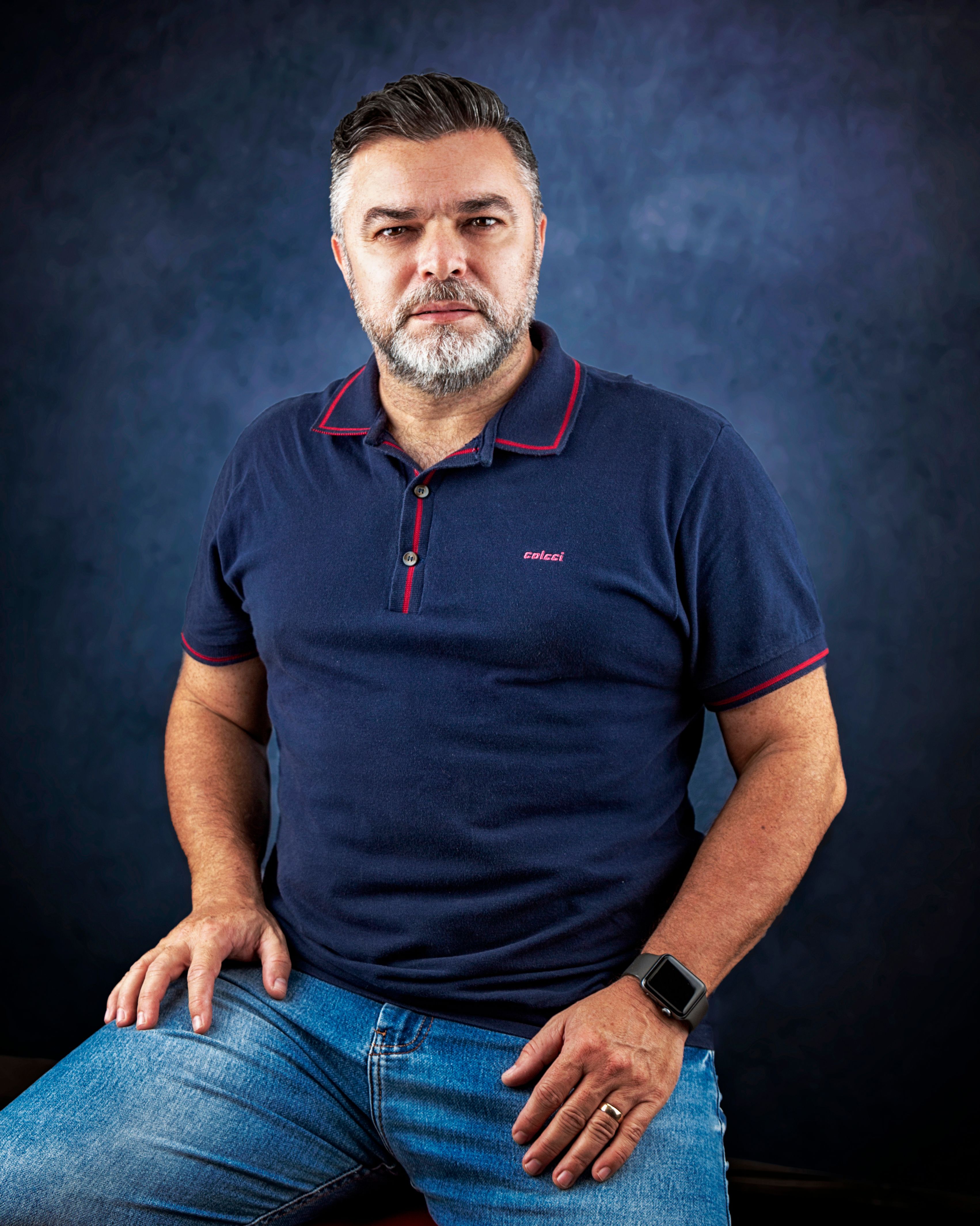 Portrait of a confident man with a beard in a studio setting wearing a navy polo shirt and jeans.