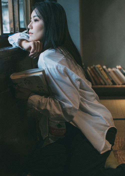 Free Asian woman in white shirt holds a book, gazing wistfully by a window indoors. Stock Photo