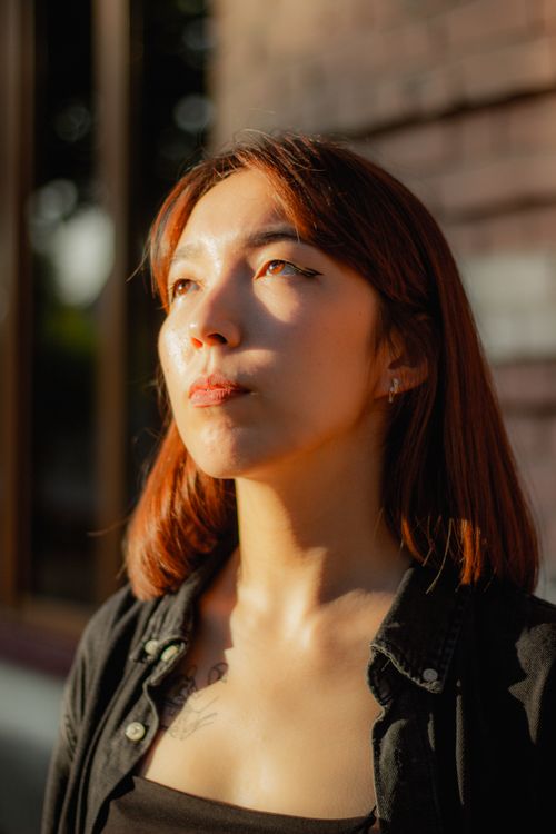 Free A young woman with red hair stands outdoors, bathed in sunlight, looking thoughtful. Stock Photo