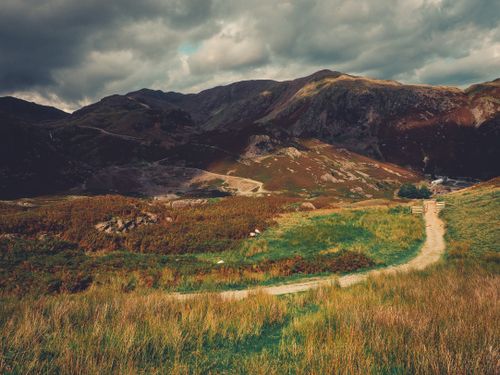 Free Beautiful rural pathway winding through lush meadow and mountains under dramatic skies. Stock Photo