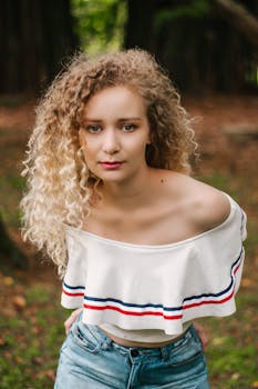 一位金发卷发女子穿着时尚露肩上衣和蓝色牛仔裤，在户外随意摆着姿势