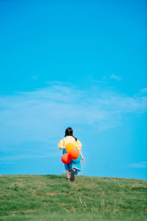 Free A young girl with colorful balloons walks up a grassy hill under a clear blue sky. Stock Photo