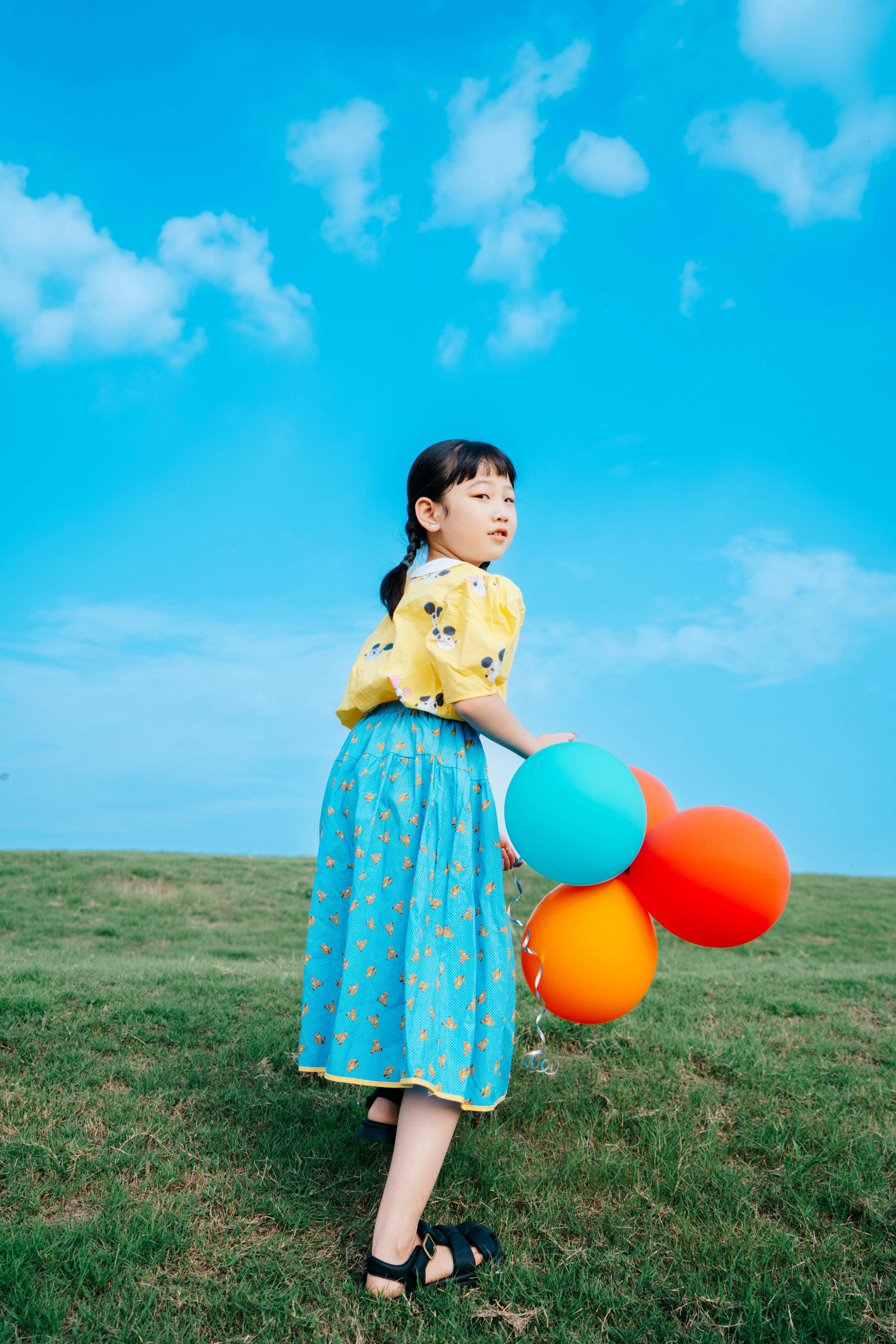 A child in colorful clothing looks back while holding balloons on a grass field under a clear blue sky.