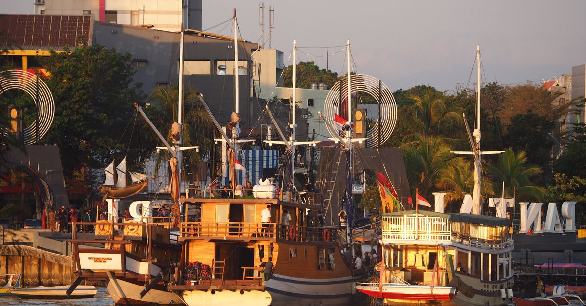 Kapal layar kayu bergaya rustic berlabuh di pelabuhan kota yang berwarna cerah saat matahari terbenam, menciptakan pemandangan perkotaan yang indah.