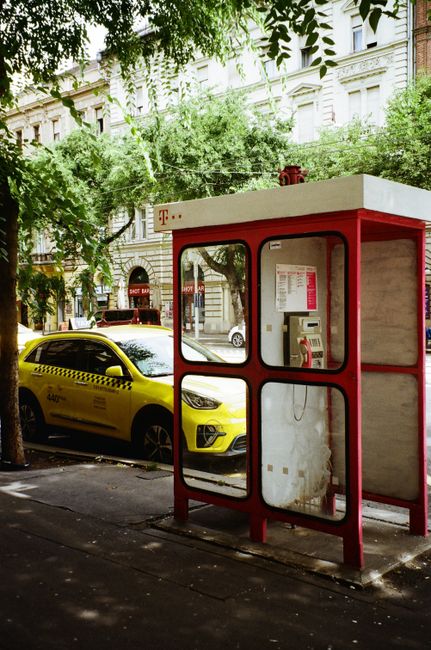 A vibrant street view in Budapest featuring a red phone booth and a yellow taxi, framed by historic architecture.