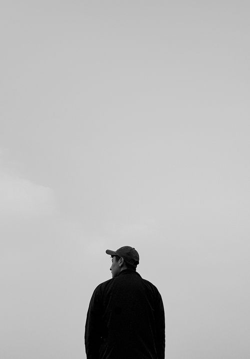 Free A man in a black jacket and baseball cap gazes up at the sky in a minimalist black and white photo. Stock Photo