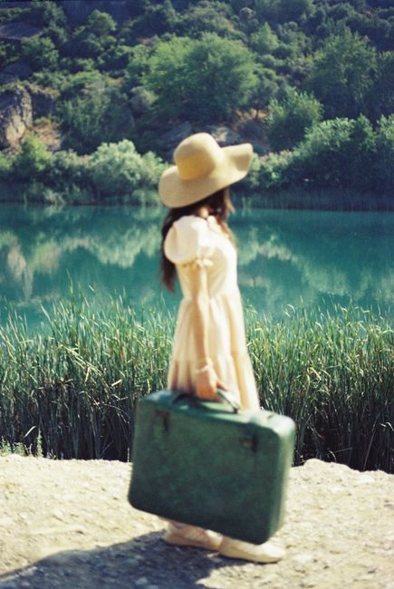 A woman in a summer dress holding a vintage suitcase by a serene lake.