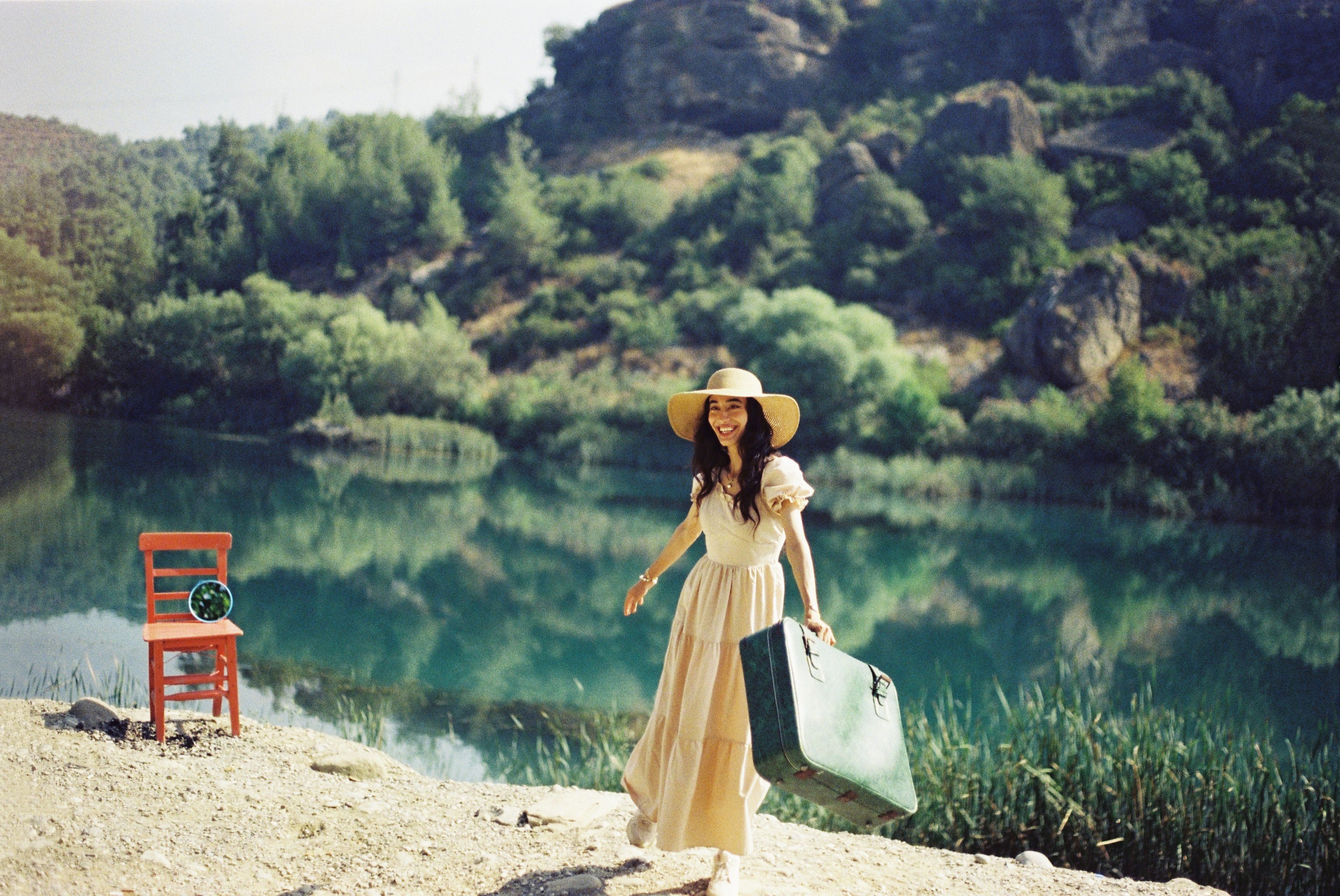 [ColoSach]-smiling-woman-in-a-hat-and-dress-by-a-scenic-lake,-holding-a-suitcase,-under-sunny-skies.