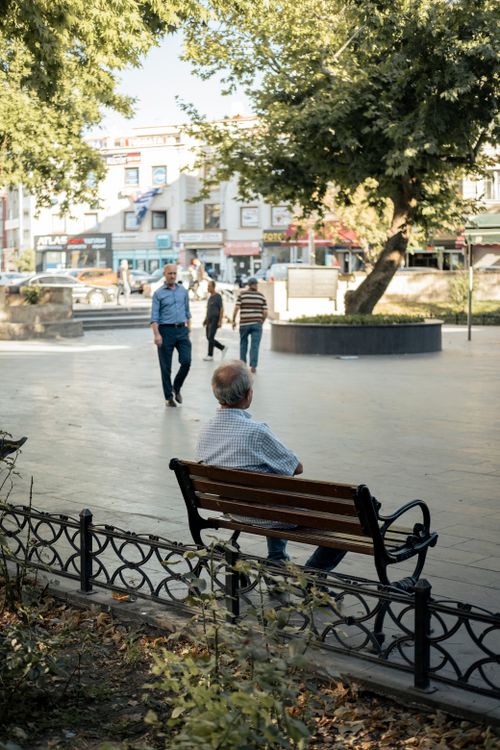 Free An elderly man sits on a bench in a city park, observing people walking by on a sunny day. Stock Photo