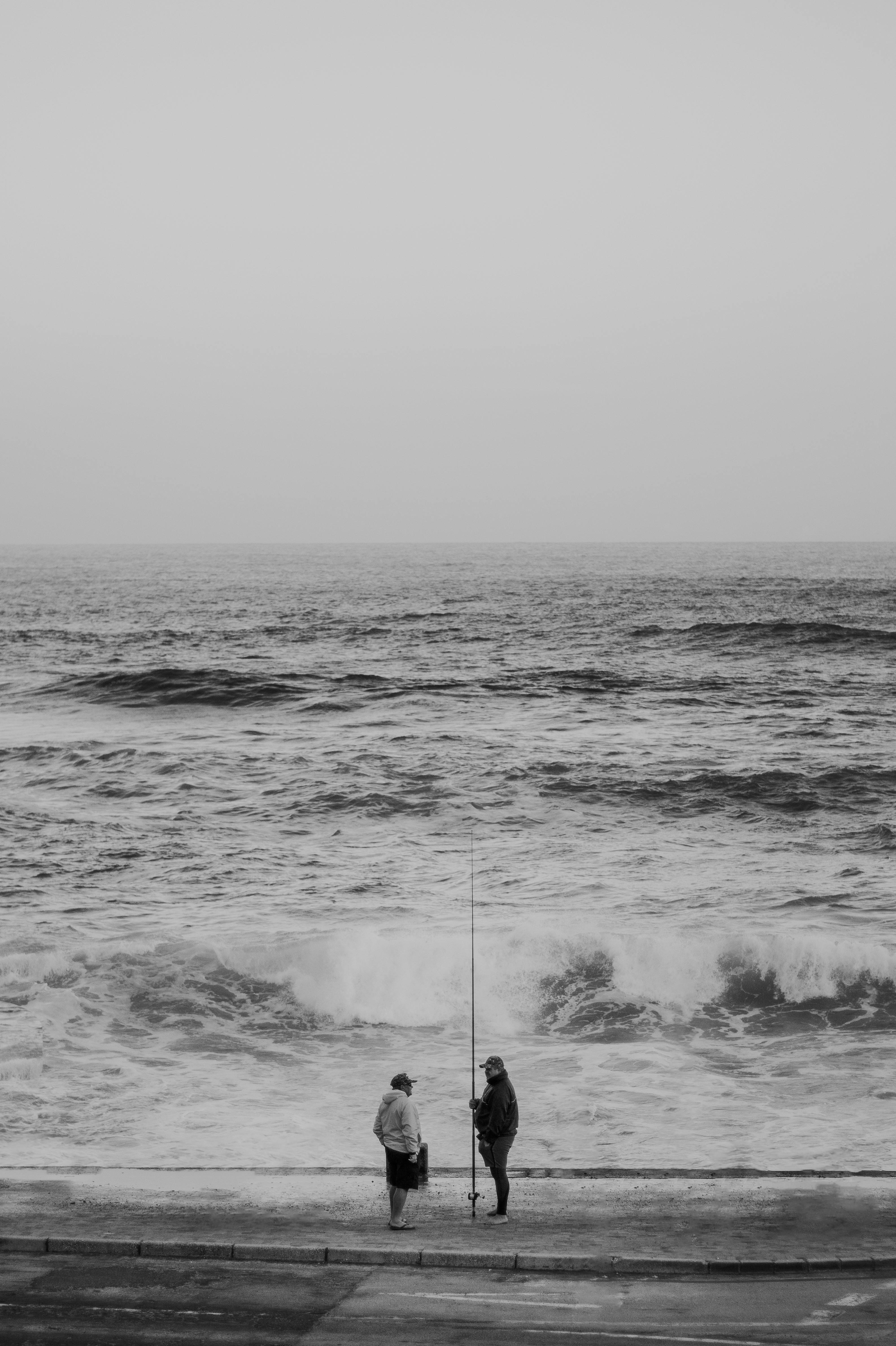Two people fishing by a rough sea on a foggy day in black and white.