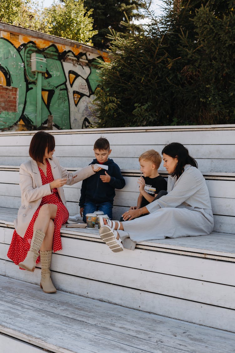 Two Women With Children Sitting On A Large White Bench