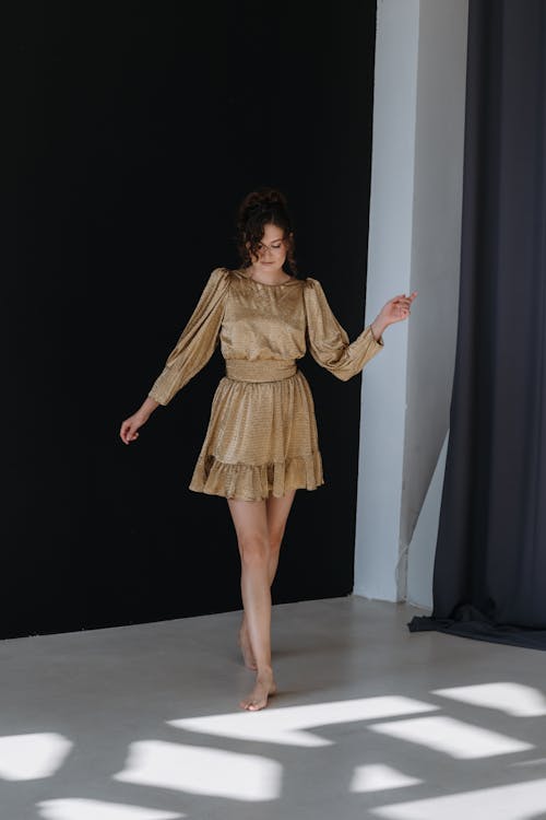 Brunette Woman in Golden Mini Dress Standing Barefoot against Black Wall