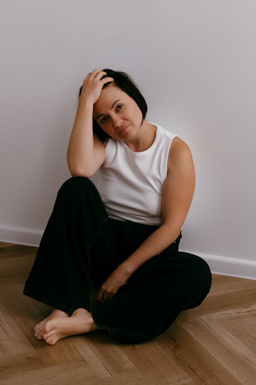 Barefoot Woman in White Tank Top and Black Pants Sitting on Wooden Floor