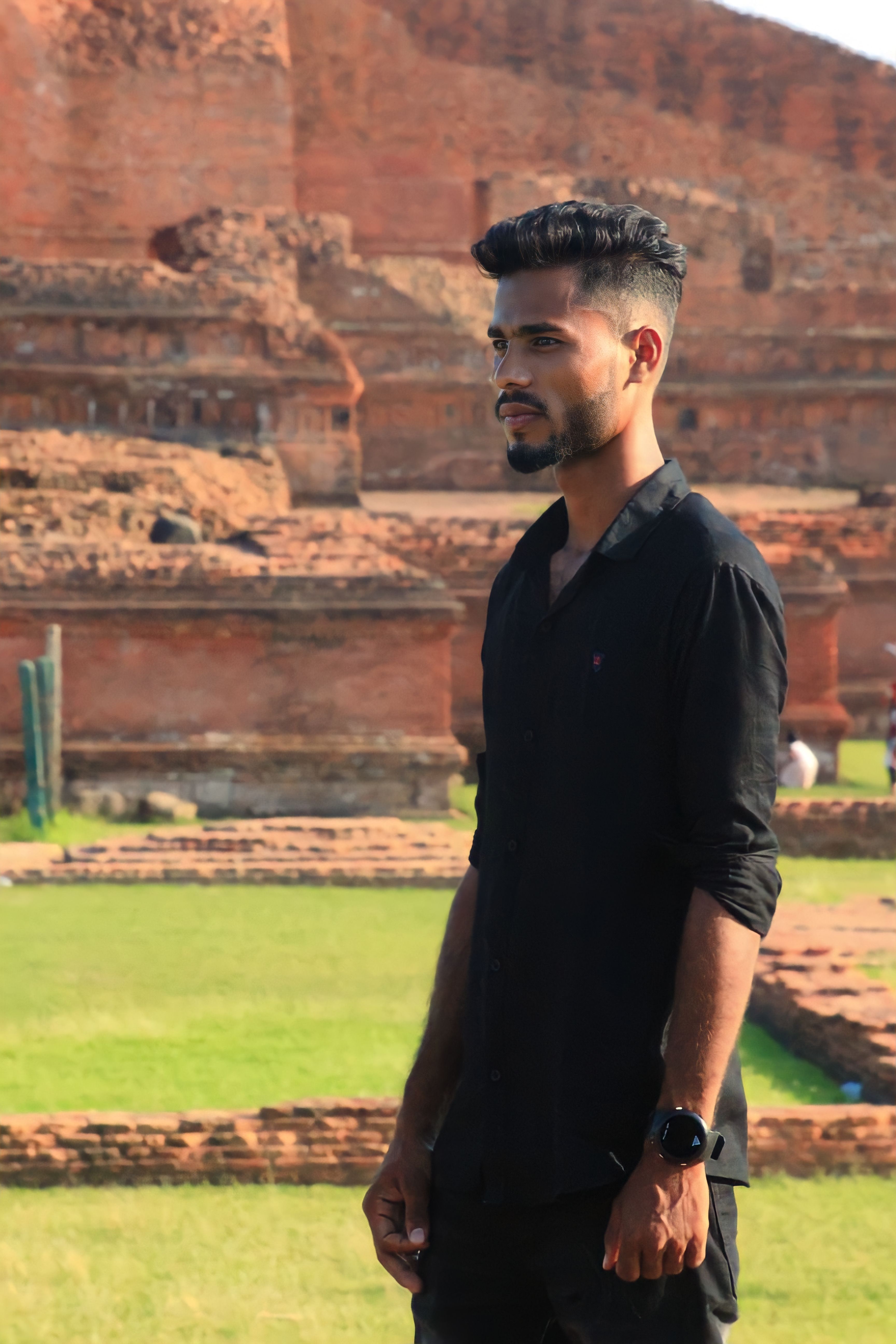 A man in a black outfit standing in front of ancient ruins, bathed in daylight.
