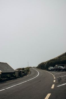 A winding road passing by parked cars and houses under an overcast sky.