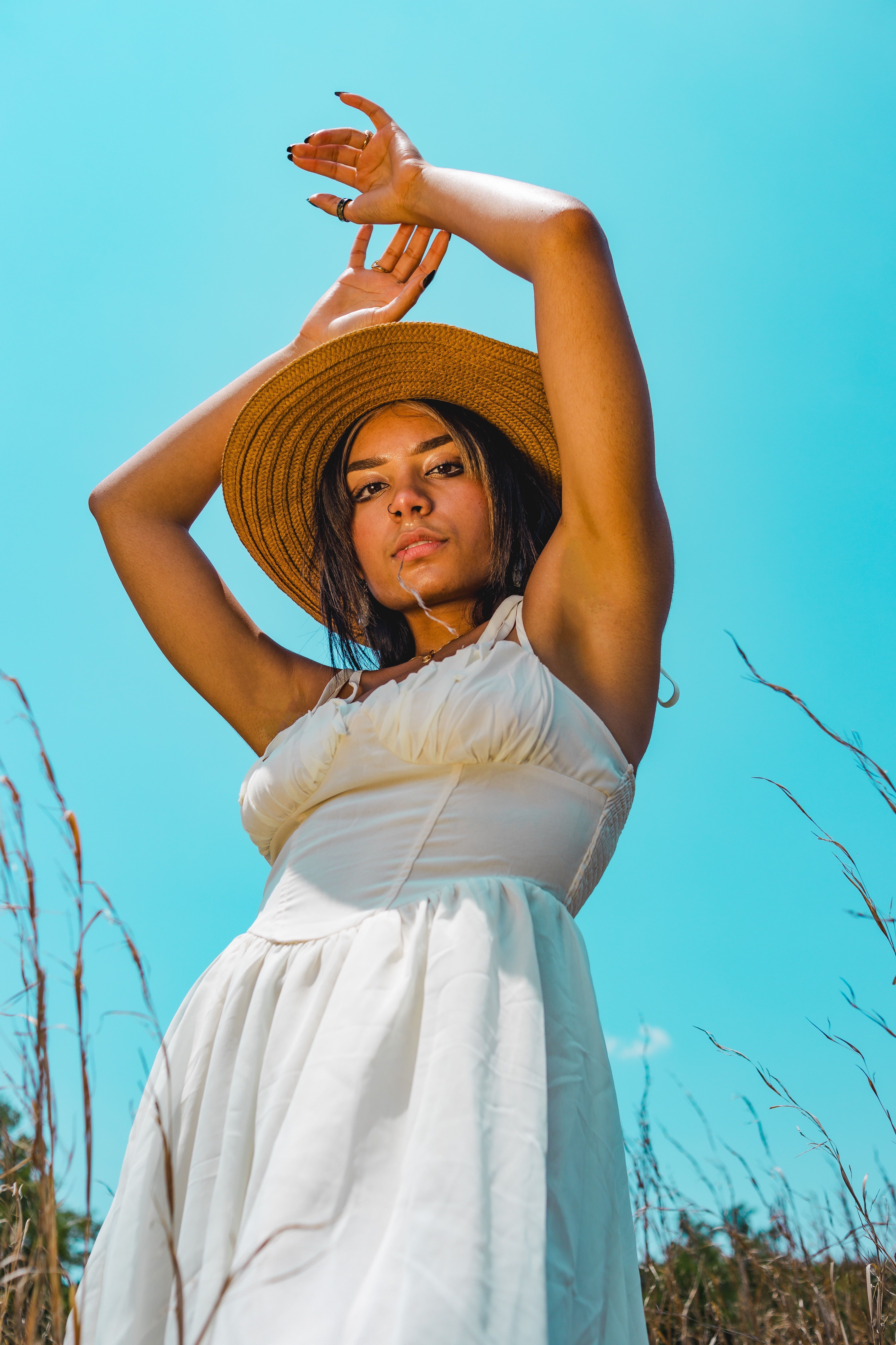 A woman poses outdoors in a white dress and straw hat against a clear blue sky.