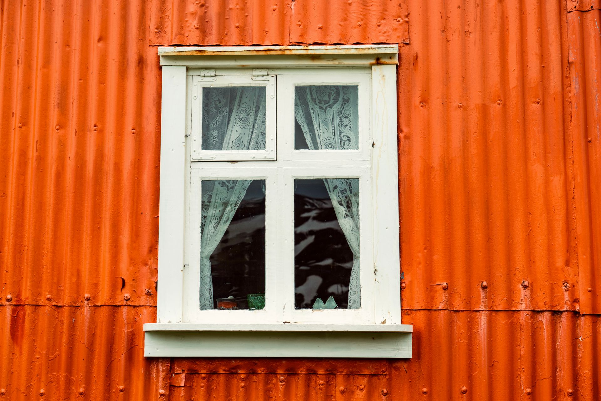 Close-up of a rustic orange metal building exterior featuring a white framed window with lace curtains.
